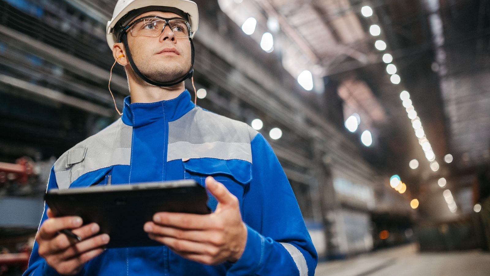 An industrial worker in protective safety gear using a tablet on a factory floor.