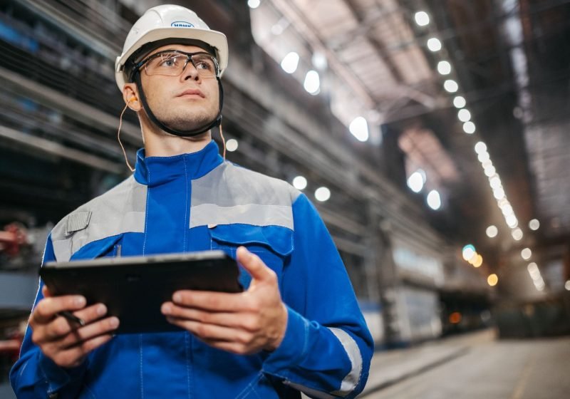An industrial worker in protective safety gear using a tablet on a factory floor.