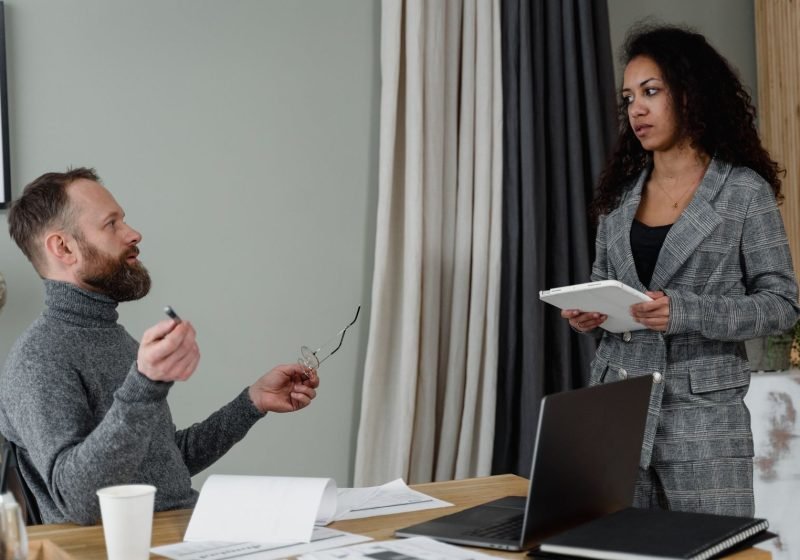A man and a woman in professional attire discussing paperwork one-to-one across a desk in a modern office.