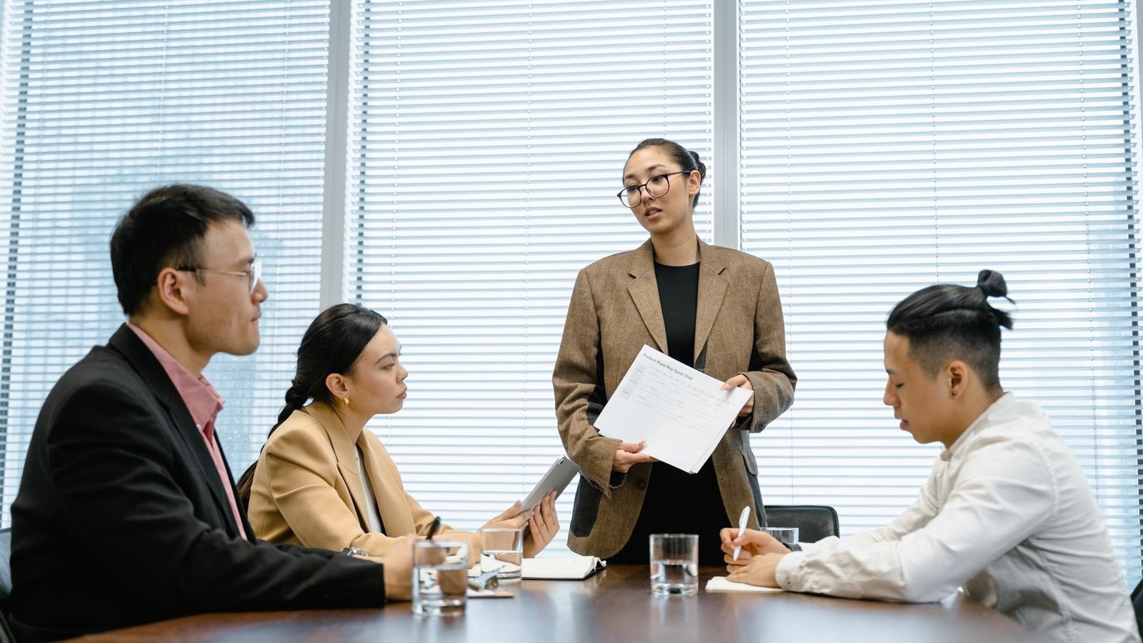 An office team in a meeting, with a woman presenting to seated colleagues in a contemporary workspace.