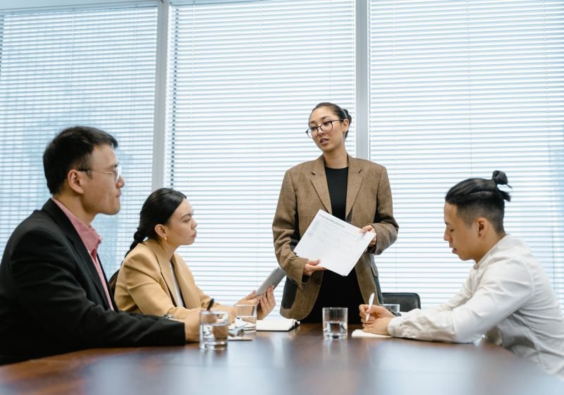 An office team in a meeting, with a woman presenting to seated colleagues in a contemporary workspace.